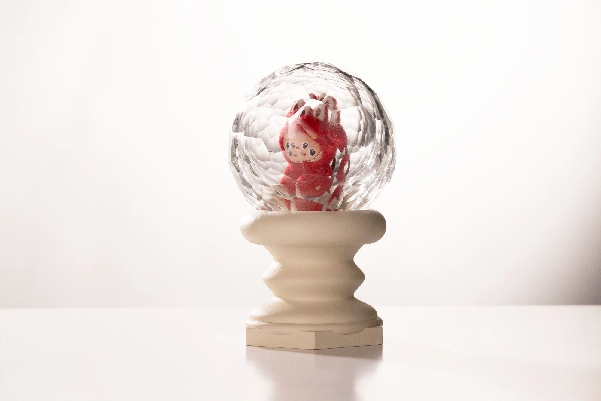 Faceted blown glass dome enclosing a red plush figure, on a cream turned pedestal, against white