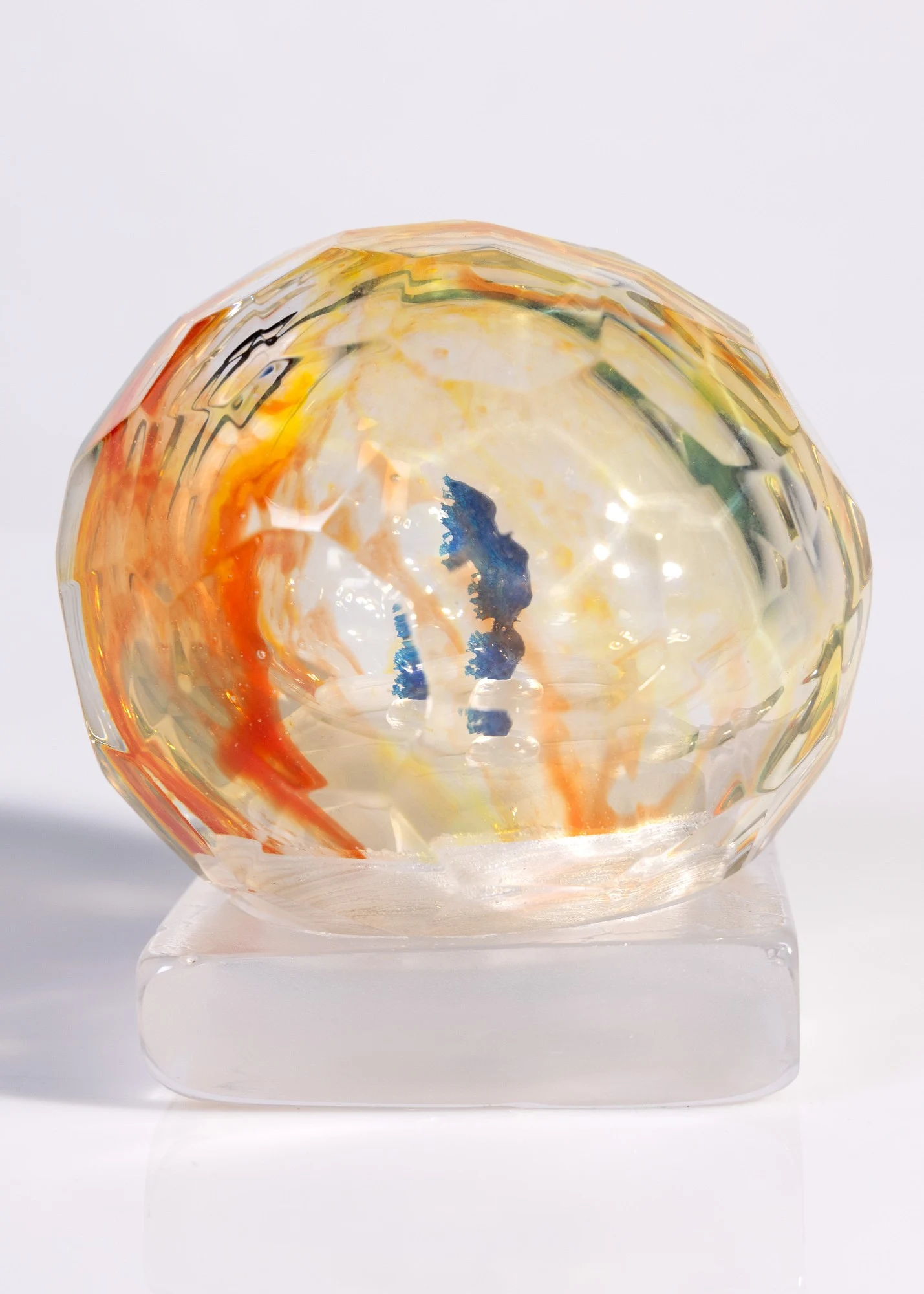 Faceted glass sphere streaked with orange and yellow, a blue pâte de verre form inside, on a frosted glass block