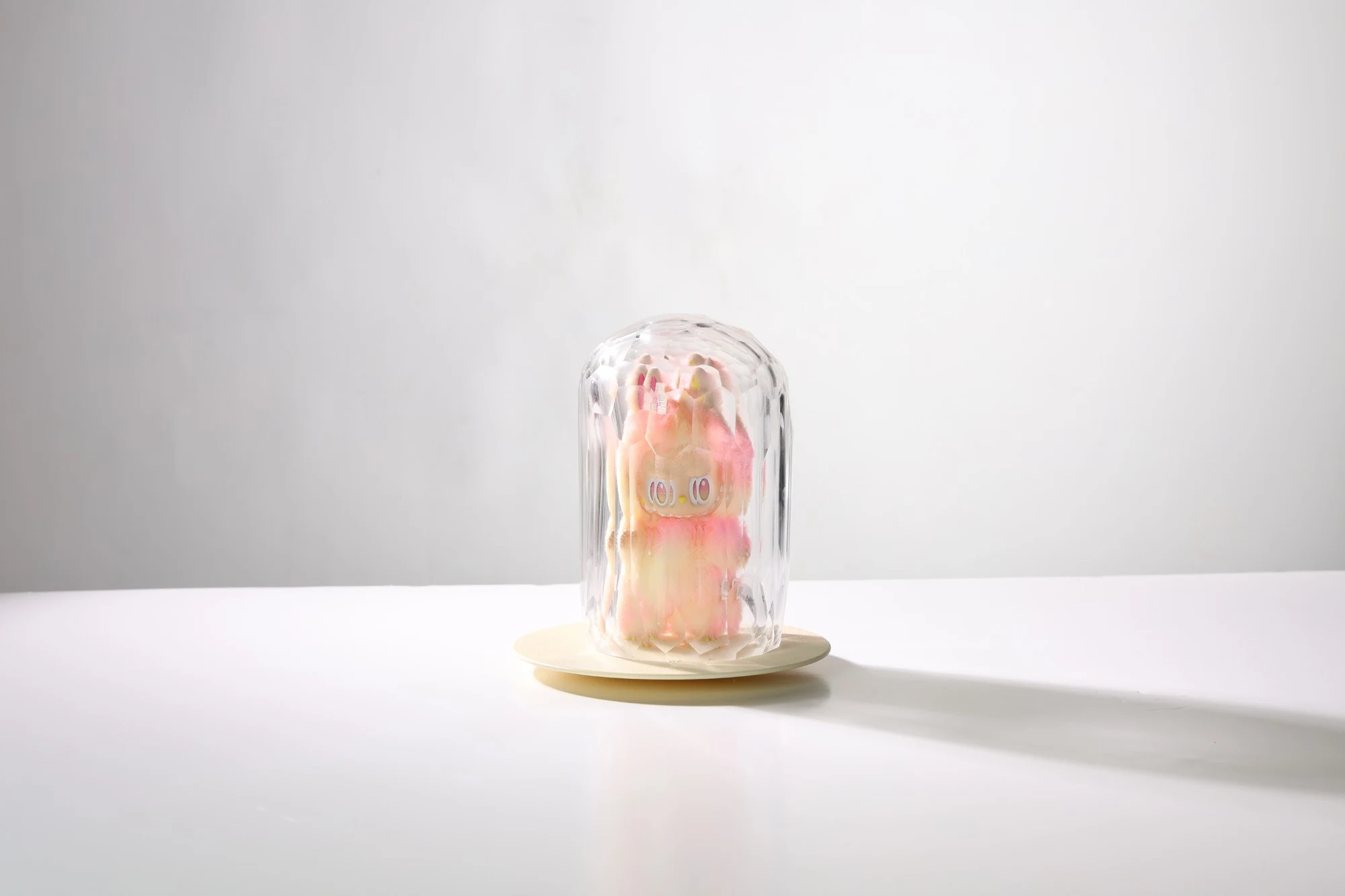 Tall faceted blown glass dome enclosing a pink and cream plush figure, on a pale round base, against white